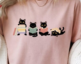 May include: A light pink t-shirt featuring a graphic print of four black cats interacting with books. One cat stands by a stack of books, two cats sit while reading open books, and one cat lies on its back with an open book resting on its head.