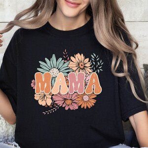 May include: Black t-shirt featuring a floral design and the word "MAMA" in colourful, retro-style lettering.