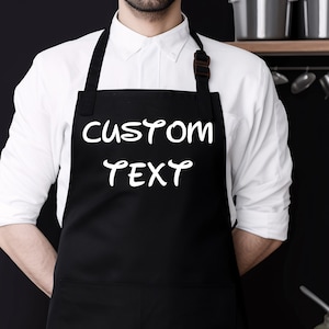 May include: A black apron with white text that says "CUSTOM TEXT". The apron is being worn by a person in a white shirt.