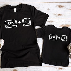 Peut inclure: Deux t-shirts noirs avec du texte blanc. Le plus grand t-shirt dit "Ctrl + C" et le plus petit dit "Ctrl + V".