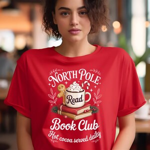 K&ouml;nnte beinhalten: Rotes T-Shirt mit einem Aufdruck mit dem Text "North Pole Book Club" und einem Design eines Lebkuchenmannes, einer Tasse hei&szlig;er Schokolade und B&uuml;chern. Der Satz "Hot cocoa served daily" ist ebenfalls enthalten.