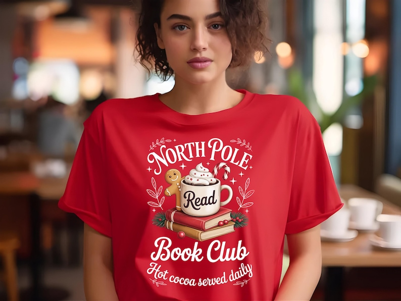 K&ouml;nnte beinhalten: Rotes T-Shirt mit einem Aufdruck mit dem Text "North Pole Book Club" und einem Design eines Lebkuchenmannes, einer Tasse hei&szlig;er Schokolade und B&uuml;chern. Der Satz "Hot cocoa served daily" ist ebenfalls enthalten.