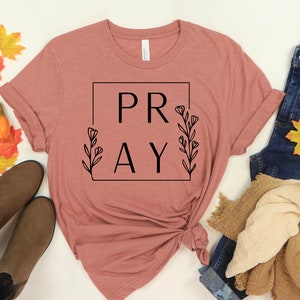 May include: A mauve t-shirt with the word "PRAY" in a black square frame with floral accents. The shirt is knotted at the bottom and is displayed on a white background with fall-themed decor.