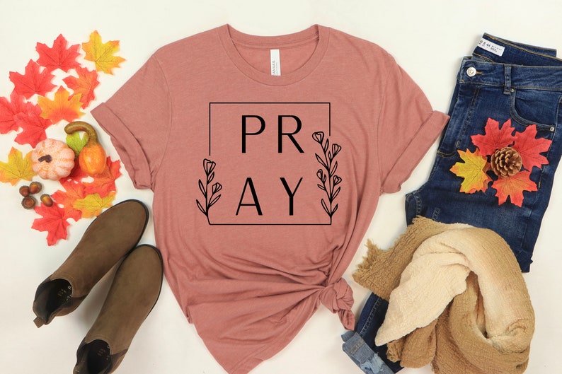 May include: A mauve t-shirt with the word "PRAY" in a black square frame with floral accents. The shirt is knotted at the bottom and is displayed on a white background with fall-themed decor.