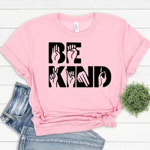 May include: Pink t-shirt with the words "BE KIND" in black lettering. The letters are formed using American Sign Language hand gestures.