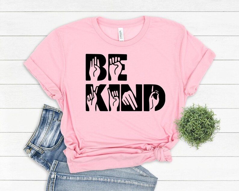 May include: Pink t-shirt with the words "BE KIND" in black lettering. The letters are formed using American Sign Language hand gestures.