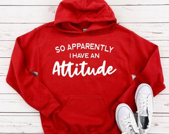 Pode incluir: Uma camisola com capuz de cor vermelha brilhante, com um bolso frontal tipo canguru e mangas compridas. O texto impresso a branco no peito diz: SO APPARENTLY I HAVE AN Attitude. A peça de vestuário é exibida com sapatilhas brancas.