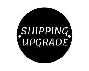 May include: A black circular sign featuring the white text SHIPPING UPGRADE in a serif font. The design includes two small white dots positioned on the left and right sides of the word SHIPPING, centred within the black circle.
