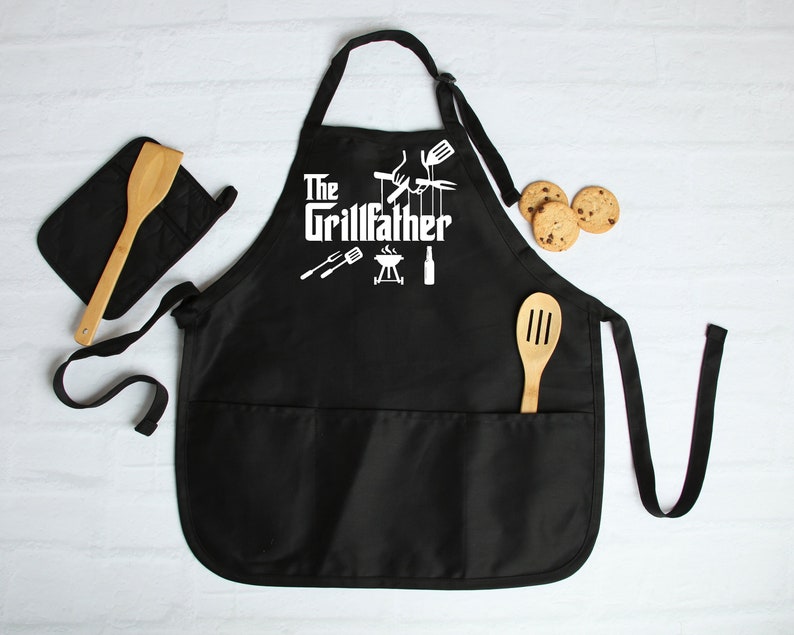 Peut inclure: Tablier de cuisine noir avec le texte "The Grillfather" en blanc, repr&eacute;sentant un barbecue, une spatule, des pinces et une bouteille en silhouette. Le tablier a deux grandes poches et des bretelles r&eacute;glables.