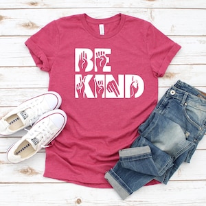 May include: A pink t-shirt with the words "BE KIND" in white lettering. The letters are formed using American Sign Language hand gestures.