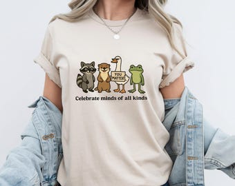 Könnte beinhalten: Ein beiges T-Shirt mit einer Grafik aus einem Waschbären, einem Otter, einer Gans mit Sonnenbrille und einem Frosch. Die Gans hält ein Schild mit der Aufschrift YOU MATTER. Unter den Tieren steht der Text Celebrate minds of all kinds.