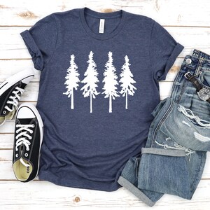 K&ouml;nnte beinhalten: Ein marineblaues T-Shirt mit einer wei&szlig;en Silhouette von vier Kiefern. Das T-Shirt liegt flach auf einer wei&szlig;en Holzoberfl&auml;che mit einem Paar schwarzen Canvas-Schuhen und einer blauen Jeans.