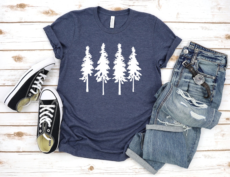 K&ouml;nnte beinhalten: Ein marineblaues T-Shirt mit einer wei&szlig;en Silhouette von vier Kiefern. Das T-Shirt liegt flach auf einer wei&szlig;en Holzoberfl&auml;che mit einem Paar schwarzen Canvas-Schuhen und einer blauen Jeans.