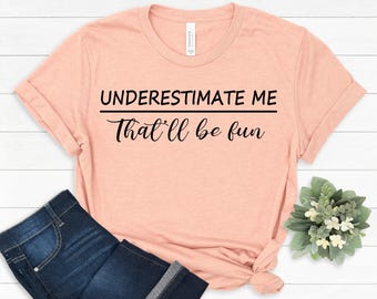 Puede incluir: Una camiseta de manga corta de color melocotón con texto negro impreso en la parte delantera que dice "UNDERESTIMATE ME" en una fuente sans-serif negrita sobre la frase "That'll be fun" en una fuente cursiva con una línea.