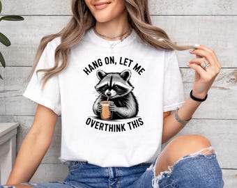 Könnte beinhalten: Ein weißes Kurzarm-T-Shirt mit einer schwarz-weißen Illustration eines Waschbären, der einen beigen Kaffeebecher mit Strohhalm hält. Der Text über dem Waschbären lautet HANG ON, LET ME und unter dem Waschbären steht OVERTHINK THIS.