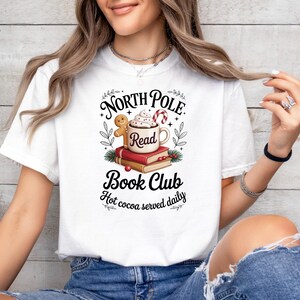 K&ouml;nnte beinhalten: Wei&szlig;es T-Shirt mit einem festlichen Aufdruck. Das Design zeigt eine Tasse hei&szlig;e Schokolade, einen Lebkuchenmann und den Text "North Pole Read Book Club". Der Satz "Hot cocoa served daily" ist ebenfalls vorhanden.