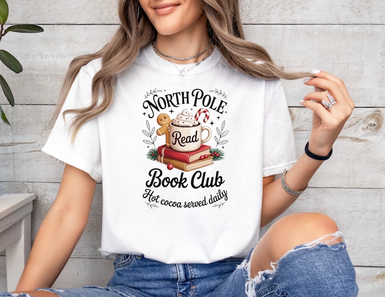 K&ouml;nnte beinhalten: Wei&szlig;es T-Shirt mit einem festlichen Aufdruck. Das Design zeigt eine Tasse hei&szlig;e Schokolade, einen Lebkuchenmann und den Text "North Pole Read Book Club". Der Satz "Hot cocoa served daily" ist ebenfalls vorhanden.