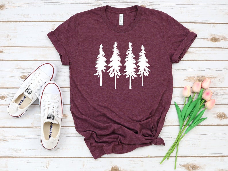 K&ouml;nnte beinhalten: Ein maroonfarbenes T-Shirt mit einer wei&szlig;en Silhouette von vier Kiefern. Das T-Shirt liegt auf einer wei&szlig;en Holzoberfl&auml;che mit einem Paar wei&szlig;en Turnschuhen und einem Strau&szlig; rosa Tulpen.