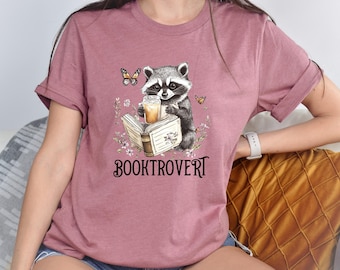 Puede incluir: Una camiseta de manga corta color malva con un gráfico de un mapache sosteniendo un vaso de café helado mientras lee un libro abierto. El diseño incluye pequeñas mariposas y detalles florales, con el texto BOOKTROVERT impreso en negro.