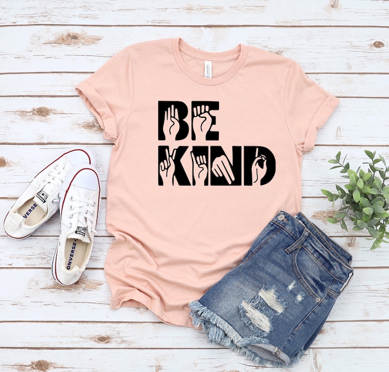May include: A pink t-shirt with the words "BE KIND" in black lettering. The letters are formed using American Sign Language hand gestures.