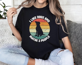 Könnte beinhalten: Ein schwarzes Kurzarm-T-Shirt mit einer kreisförmigen Grafik, die einen Retro-Sonnenuntergang in orangefarbenen, gelben und grünen Streifen zeigt. Eine schwarze Hundesilhouette sitzt in der Mitte, umgeben vom Text I LIKE DOGS AND MAYBE 3 PEOPLE.