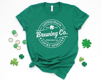 Puede incluir: Una camiseta verde de manga corta con un gráfico circular blanco que dice "LEPRECHAUN Brewing Co. EST. 1993 PREMIUM LUCKY SPIRITS", con un icono de trébol de cuatro hojas en la parte superior y pequeños detalles de tréboles.