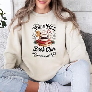 K&ouml;nnte beinhalten: Cremefarbenes Sweatshirt mit einem Aufdruck &bdquo;North Pole Book Club&ldquo;. Das Design umfasst eine Tasse hei&szlig;e Schokolade, einen Lebkuchenmann, eine Zuckerstange und B&uuml;cher. Der Satz &bdquo;Hot cocoa served daily&ldquo; ist ebenfalls enthalten.