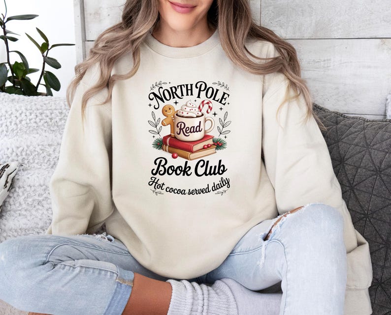 K&ouml;nnte beinhalten: Cremefarbenes Sweatshirt mit einem Aufdruck &bdquo;North Pole Book Club&ldquo;. Das Design umfasst eine Tasse hei&szlig;e Schokolade, einen Lebkuchenmann, eine Zuckerstange und B&uuml;cher. Der Satz &bdquo;Hot cocoa served daily&ldquo; ist ebenfalls enthalten.