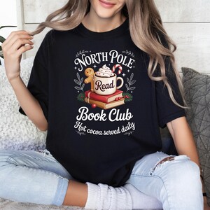 K&ouml;nnte beinhalten: Schwarzes T-Shirt mit dem Aufdruck "North Pole Book Club". Das Design zeigt eine Tasse hei&szlig;e Schokolade, B&uuml;cher, einen Lebkuchenmann und Zuckerstangen. Der Text "Hot cocoa served daily" ist ebenfalls enthalten.