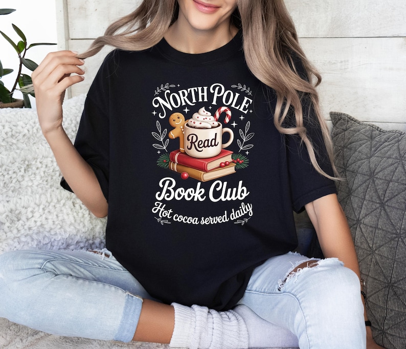 K&ouml;nnte beinhalten: Schwarzes T-Shirt mit dem Aufdruck "North Pole Book Club". Das Design zeigt eine Tasse hei&szlig;e Schokolade, B&uuml;cher, einen Lebkuchenmann und Zuckerstangen. Der Text "Hot cocoa served daily" ist ebenfalls enthalten.