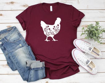 May include: A maroon short-sleeved t-shirt featuring a white silhouette of a chicken filled with a floral pattern. The shirt is displayed alongside a pair of blue denim jeans and white canvas sneakers, showcasing a casual ensemble.