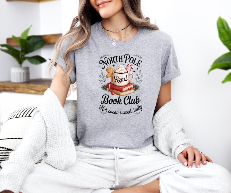 K&ouml;nnte beinhalten: Ein meliertes graues T-Shirt mit dem Aufdruck "North Pole Read Book Club". Das Design zeigt eine Tasse hei&szlig;e Schokolade, B&uuml;cher, einen Lebkuchenmann und Zuckerstangen. Der Satz "Hot cocoa served daily" ist ebenfalls enthalten.