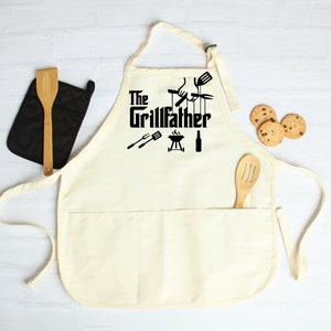 Peut inclure: Un tablier en coton blanc avec un grand motif noir qui dit "The Grillfather" avec un barbecue, une bouteille de bi&egrave;re et des outils de grillade. Le tablier a une grande poche sur le devant.