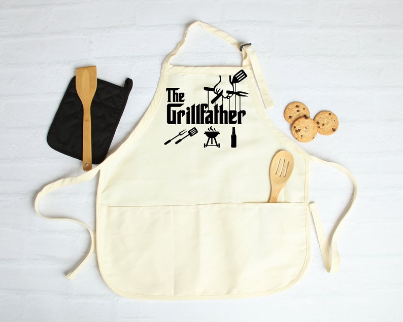 Peut inclure: Un tablier en coton blanc avec un grand motif noir qui dit "The Grillfather" avec un barbecue, une bouteille de bi&egrave;re et des outils de grillade. Le tablier a une grande poche sur le devant.