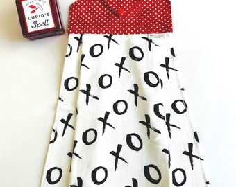 May include: A white kitchen towel with a red and white polka dot hanging loop. The towel has a black and white XOXO pattern. A small red bottle with a heart-shaped stopper and the text "Cupid's Spell" is in the background.