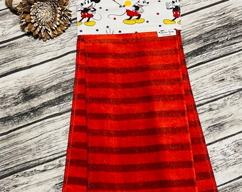 May include: A hanging kitchen towel featuring a red and orange striped design. The top portion has a white background with Mickey Mouse illustrations and red stars. The towel is made of a soft, absorbent material.