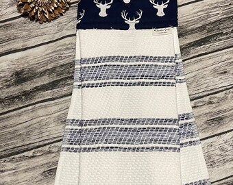 May include: A white and navy blue kitchen towel with a deer head pattern on the top flap. The towel has a striped pattern in shades of blue and white. The towel is hanging on a wooden surface.