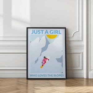 May include: A framed vertical poster featuring a stylized illustration of a snowboarder in a pink suit on a snowy mountain. The text at the top reads JUST A GIRL and the text at the bottom reads WHO LOVES THE SLOPES in light blue lettering.