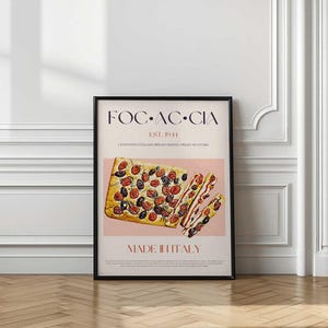 May include: A framed poster featuring a rectangular focaccia bread topped with tomatoes and olives. The poster has the text "FOC-AC-CIA EST. 1941 MADE IN ITALY". The bread is sliced, showing the interior. The background is a light pink color.