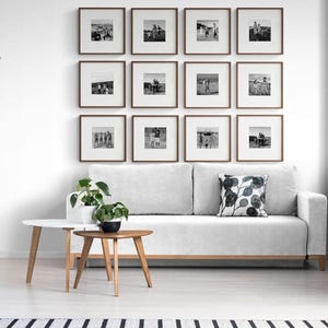 May include: A modern living room with a white sofa, two wooden side tables, and a wall-mounted grid of framed black and white photographs. The room is decorated with plants and a patterned pillow. The wall art is from We Frame Art.