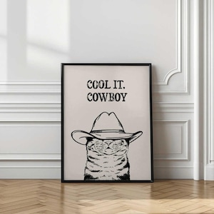 May include: Black framed art print featuring a cat wearing a cowboy hat. The black and white illustration has the text "COOL IT. COWBOY" above the cat's head. The artwork is displayed on a light beige background.