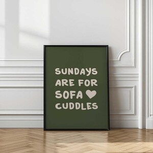 May include: A framed art print with the text "SUNDAYS ARE FOR SOFA CUDDLES" in white on a dark green background. The print is in a black frame and is displayed against a white wall with decorative molding and a wooden floor.