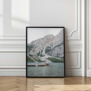 May include: A framed wall art print featuring a landscape photograph of a calm lake with several small wooden rowboats floating near the shore, set against a backdrop of towering, rugged grey mountains and a dense forest of evergreen trees.