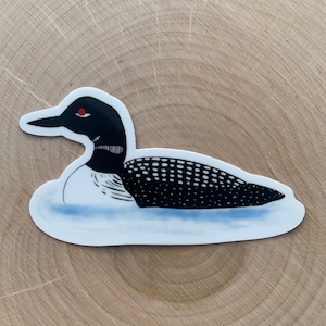 May include: A black and white loon sticker with red eyes, swimming on a blue and white water background.