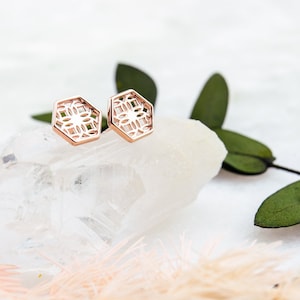 May include: A pair of rose gold earrings with a geometric design. The earrings are hexagonal in shape and feature a delicate, intricate pattern.