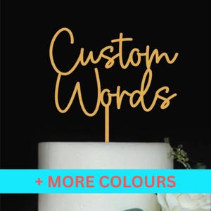 May include: A gold cake topper with the words "Custom Words" in a cursive font. The topper is placed on a white cake. A blue banner at the bottom reads "+ MORE COLOURS". The background is black.