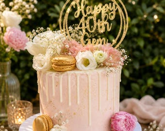 Op de afbeelding: Een cilindrische taart met lichtroze glazuur en witte druipende glazuur, versierd met witte en roze bloemen, gouden macarons en een ronde, goudkleurige glittertaarttopper met de tekst Happy 40th Gem in een cursief lettertype.