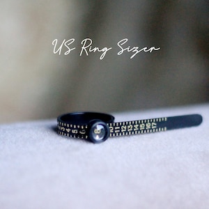 May include: A black plastic ring sizer with gold numbers printed on it. The sizer is marked with US ring sizes from 4 to 17. The text "US Ring Sizer" is written in white on a blurred background.