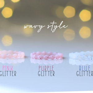 Pode incluir: Tr&ecirc;s an&eacute;is de silicone com um design ondulado. Os an&eacute;is s&atilde;o rosa com glitter rosa, roxo com glitter roxo e branco com glitter azul. O texto "wavy style" est&aacute; escrito acima dos an&eacute;is.
