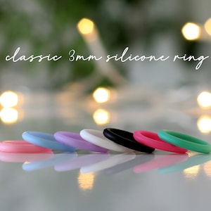 May include: A set of eight silicone rings in various colors, including pink, blue, lavender, white, black, and green. The rings are 3mm wide and are arranged in a row on a reflective surface. The text "classic 3mm silicone ring" is written above the rings.